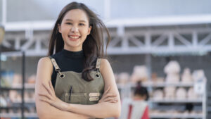 Asia female ceramist with apron hand confident chest in her workshop clay sculpture studio with positive smiling warm welcome ready to start new factory ceramic workshop with new collection of work